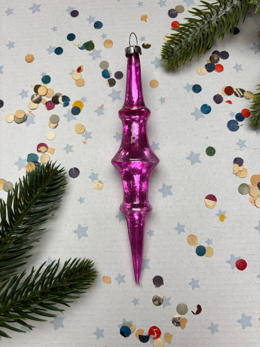 A pink glass ornament with a pointed end is surrounded by colorful confetti and pine branches on a white background.