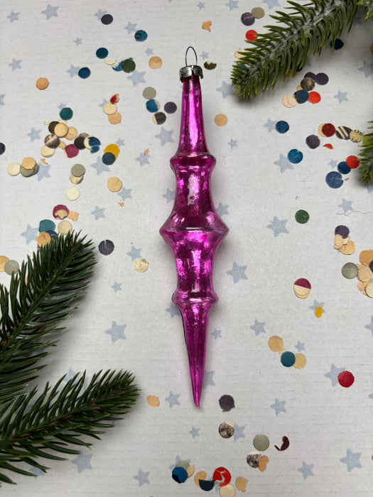A pink glass ornament with a pointed end is surrounded by colorful confetti and pine branches on a white background.