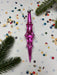 A pink glass ornament with a pointed end is surrounded by colorful confetti and pine branches on a white background.