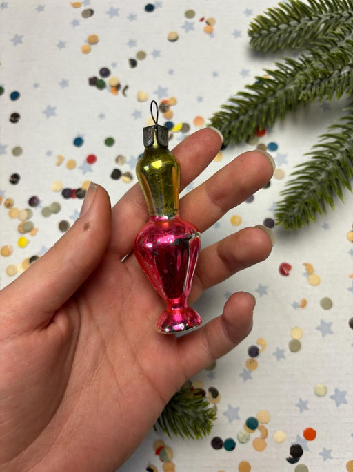 A hand holding a small, colorful glass ornament against a background of confetti and a pine tree branch.