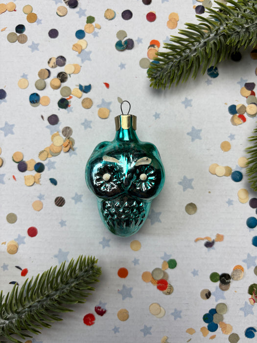 A turquoise glass ornament with a gold-colored stem is placed on a white background, surrounded by scattered confetti and green pine branches.