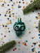A turquoise glass ornament with a gold-colored stem is placed on a white background, surrounded by scattered confetti and green pine branches.
