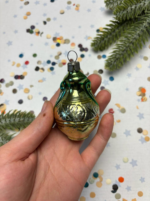 A hand holding a small, ornate green ornament with a gold-colored base, set against a background of colorful confetti and pine branches.
