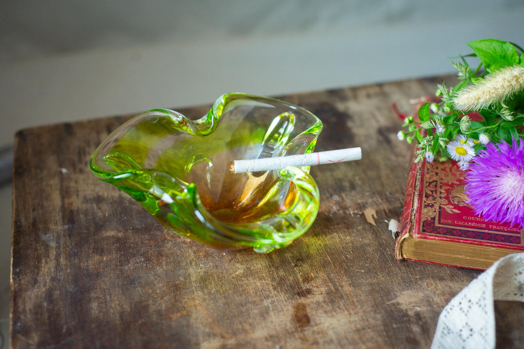 A green glass ashtray with a lit cigarette rests on a wooden surface, accompanied by a red book and a bouquet of purple flowers.