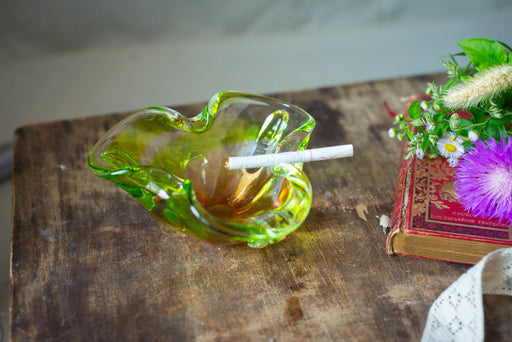 A green glass ashtray with a lit cigarette rests on a wooden surface, accompanied by a red book and a bouquet of purple flowers.