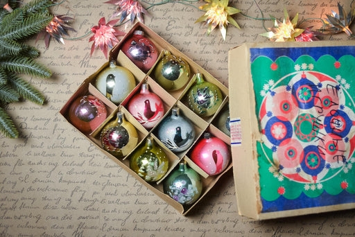 A box of colorful Christmas ornaments, including various shapes and sizes, is placed on a surface with a handwritten note and some decorative elements.
