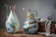 two colorful glass pitchers, one with a long handle and the other with a shorter handle, placed on a wooden surface alongside a bouquet of flowers and some pearls.