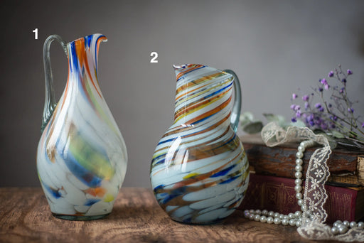 two colorful glass pitchers, one with a long handle and the other with a shorter handle, placed on a wooden surface alongside a bouquet of flowers and some pearls.