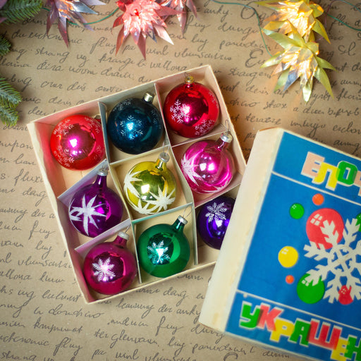 A box of colorful Christmas ornaments, including red, blue, green, and yellow balls, is placed on a surface with handwritten text and decorative paper.