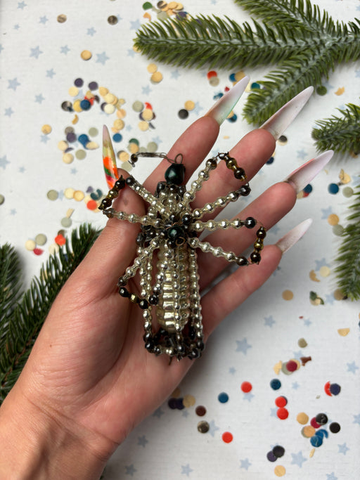 A hand holding a beaded spider-shaped ornament against a background of confetti and pine branches.