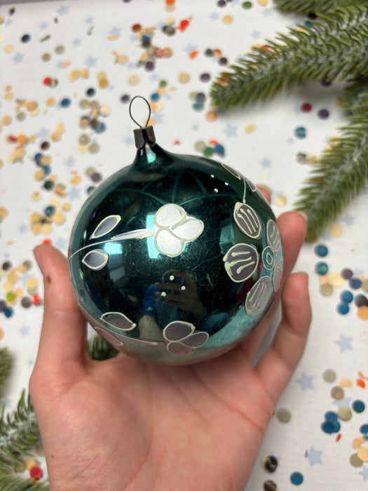 A hand holding a dark green ornament with a floral design, against a background of colorful confetti and a pine tree branch.