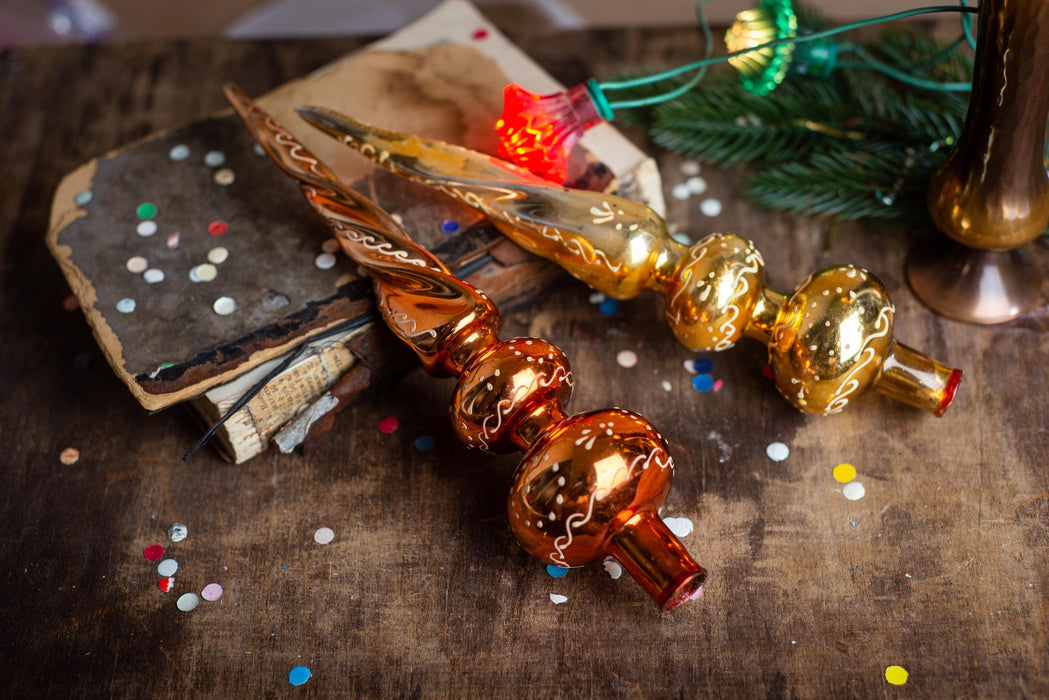 two ornate glass ornaments, one with a red light at the top and the other with a gold light, placed on a wooden surface. There are also scattered confetti around the ornaments, and a book and a candle are visible in the background.