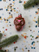 A pink Christmas ornament with a star design sits on a background of colorful confetti and pine branches.