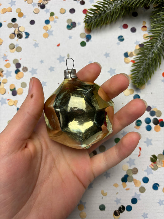 A hand holding a shiny, metallic ornament against a background of colorful confetti and a pine branch.