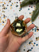 A hand holding a shiny, metallic ornament against a background of colorful confetti and a pine branch.