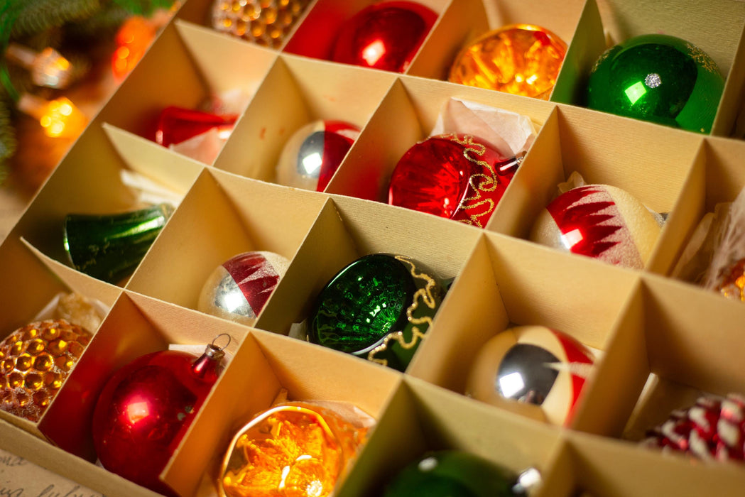 A close-up view of a collection of colorful Christmas ornaments arranged in a box, with a blurred background suggesting a festive setting.