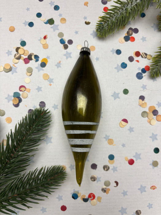 A green ornament with white stripes is placed on a white background, surrounded by colorful confetti and a sprig of pine.