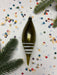 A green ornament with white stripes is placed on a white background, surrounded by colorful confetti and a sprig of pine.
