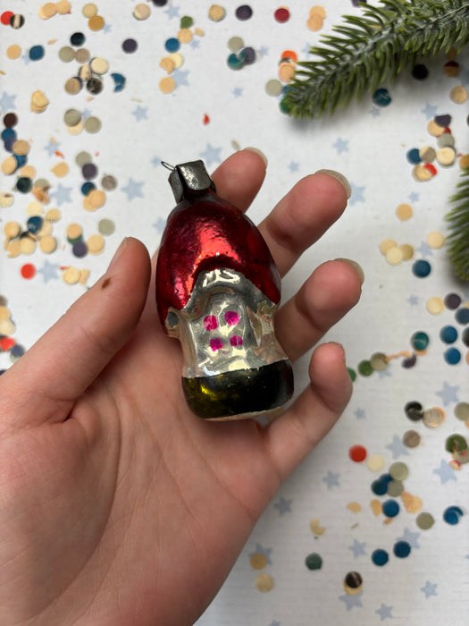 A hand holding a small, colorful ornament against a background of confetti and a pine branch.