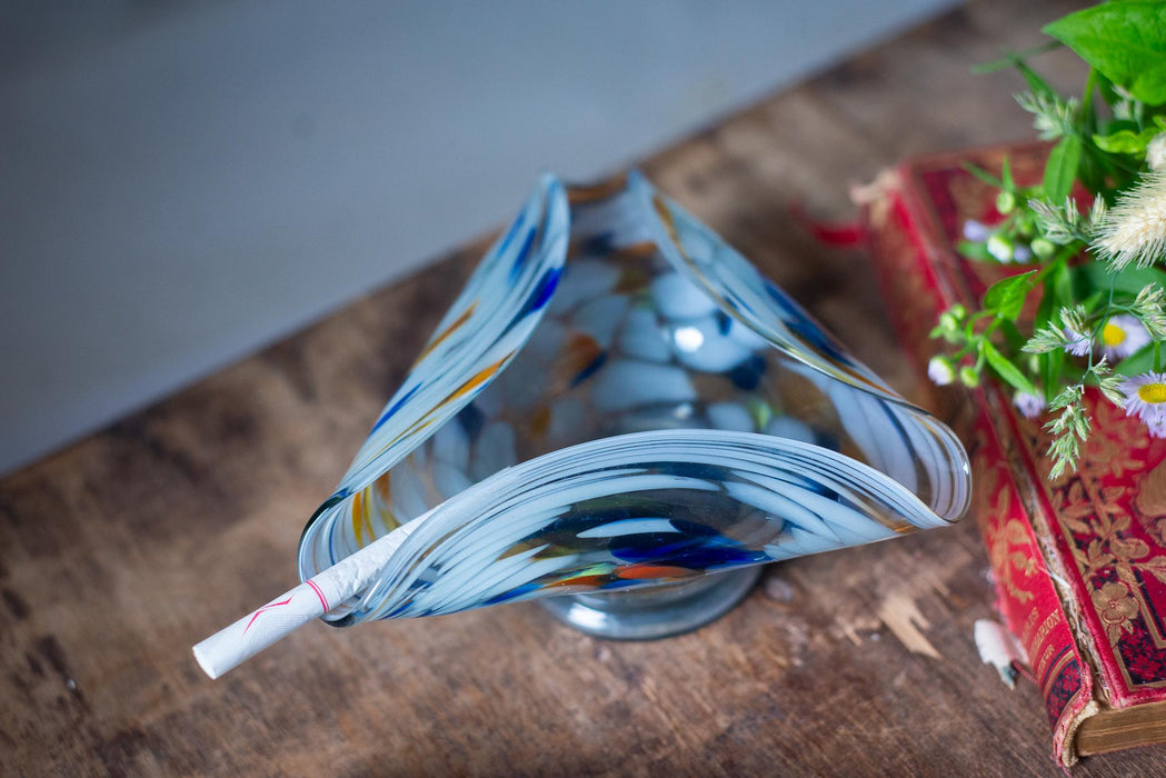 A blue and white glass ashtray with a lit cigarette rests on a wooden surface, accompanied by a red and gold book and a bouquet of flowers.