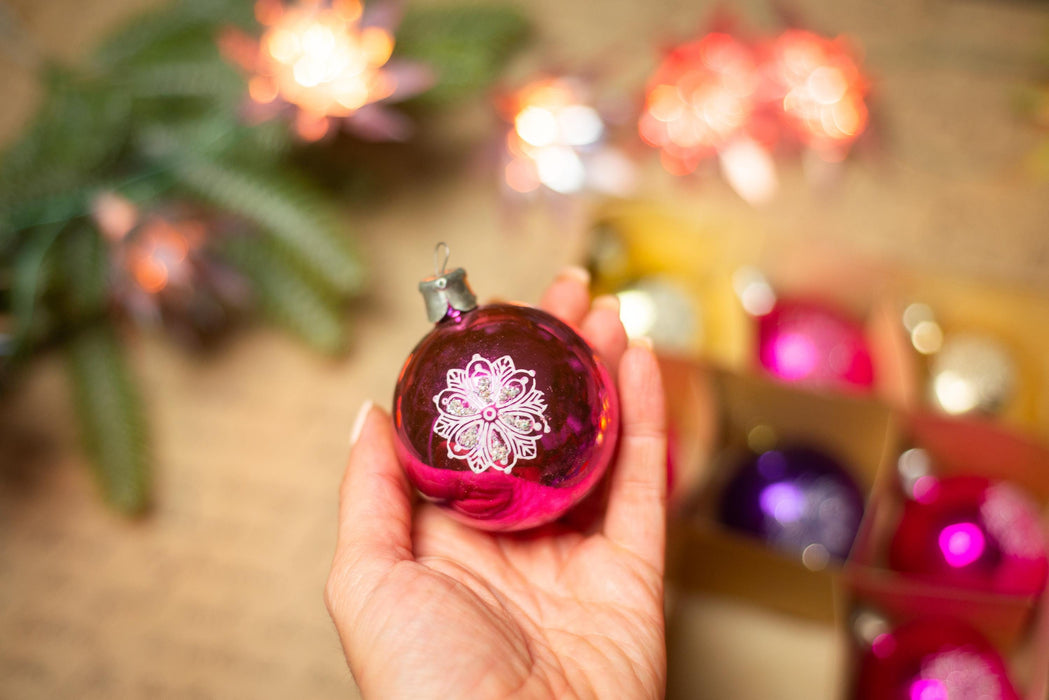A hand holding a pink ornament with a white flower design, surrounded by other colorful Christmas ornaments.