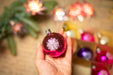 A hand holding a pink ornament with a white flower design, surrounded by other colorful Christmas ornaments.