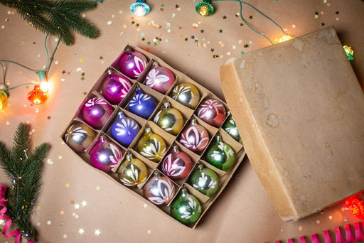 A cardboard box filled with colorful Christmas ornaments, surrounded by festive decorations such as lights, greenery, and confetti.