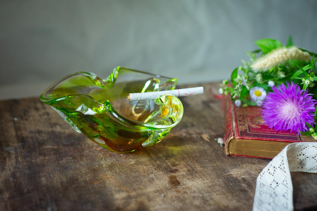A green glass ashtray with a lit cigarette rests on a wooden surface, accompanied by a bouquet of purple flowers and an old book.