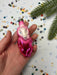 A hand holding a pink and purple ornament with a whimsical design, surrounded by confetti and a green pine branch.
