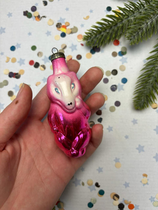 A hand holding a pink and purple ornament with a whimsical design, surrounded by confetti and a green pine branch.