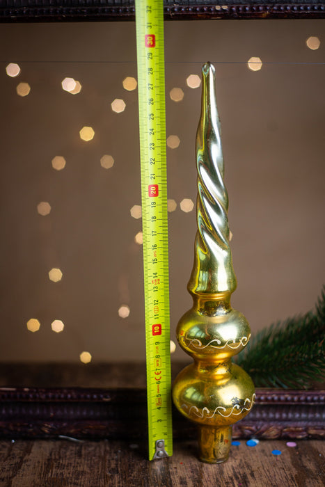 A golden Christmas ornament with a spiral design is placed next to a yellow measuring tape on a wooden surface, with a blurred background of lights.