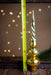 A golden Christmas ornament with a spiral design is placed next to a yellow measuring tape on a wooden surface, with a blurred background of lights.