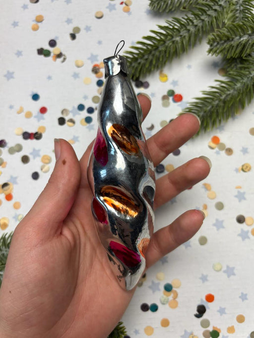 A hand holding a silver ornament with colorful designs against a background of confetti and a pine tree branch.