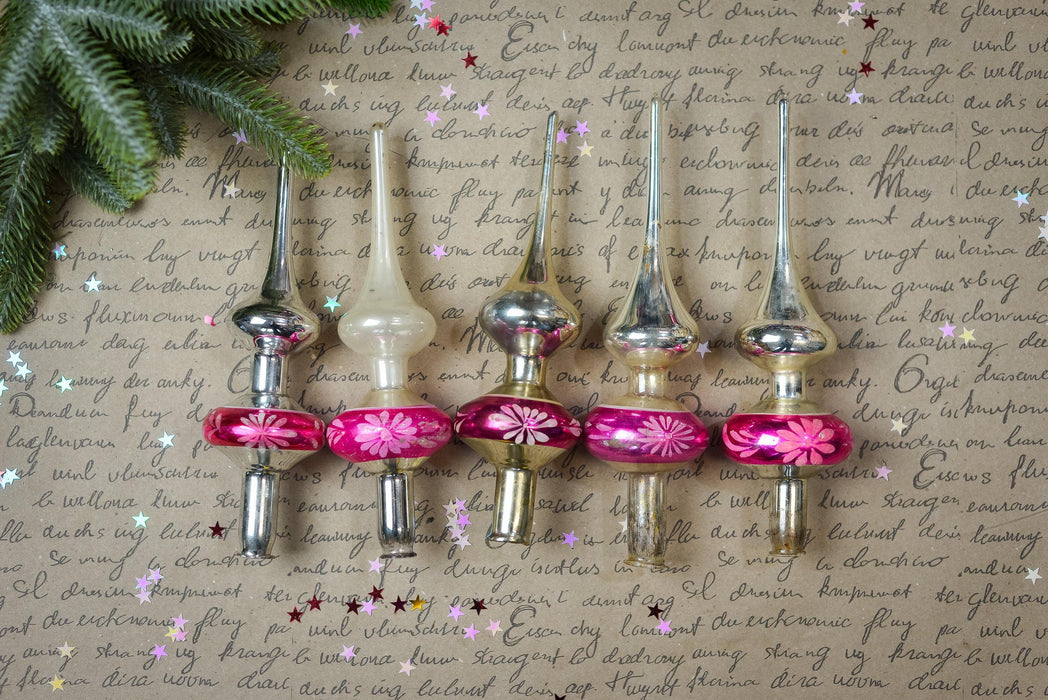 a collection of Christmas ornaments, including glass and metal pieces, arranged on a textured surface with a hint of greenery. The ornaments have a pink and silver color scheme and are placed in a row, creating a visually appealing display.