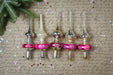 a collection of Christmas ornaments, including glass and metal pieces, arranged on a textured surface with a hint of greenery. The ornaments have a pink and silver color scheme and are placed in a row, creating a visually appealing display.