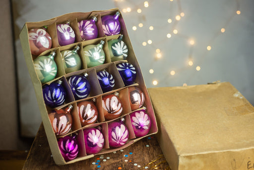 A cardboard box filled with colorful Christmas ornaments, including purple, blue, and pink ornaments, is placed on a wooden surface with twinkling lights in the background.
