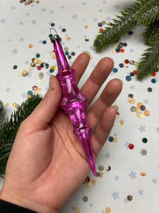 A hand holding a pink ornament against a background of confetti and pine branches.