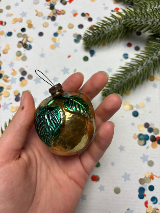 A hand holding a decorative ornament with green leaves, against a background of colorful confetti and a pine tree branch.