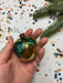 A hand holding a decorative ornament with green leaves, against a background of colorful confetti and a pine tree branch.
