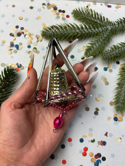 A hand holding a small, ornate ornament with a bell-like shape and a pink pendant, surrounded by Christmas tree branches and confetti.