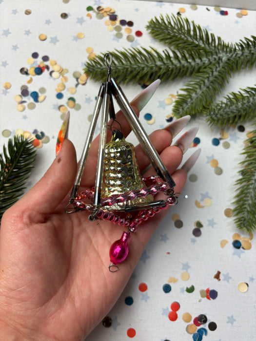A hand holding a small, ornate ornament with a bell-like shape and a pink pendant, surrounded by Christmas tree branches and confetti.