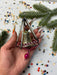 A hand holding a small, ornate ornament with a bell-like shape and a pink pendant, surrounded by Christmas tree branches and confetti.