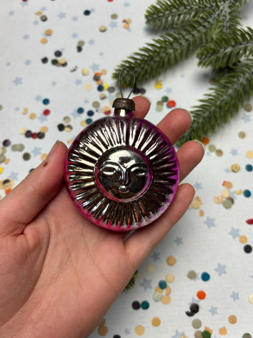 A hand holding a round ornament with a sun design, surrounded by colorful confetti.
