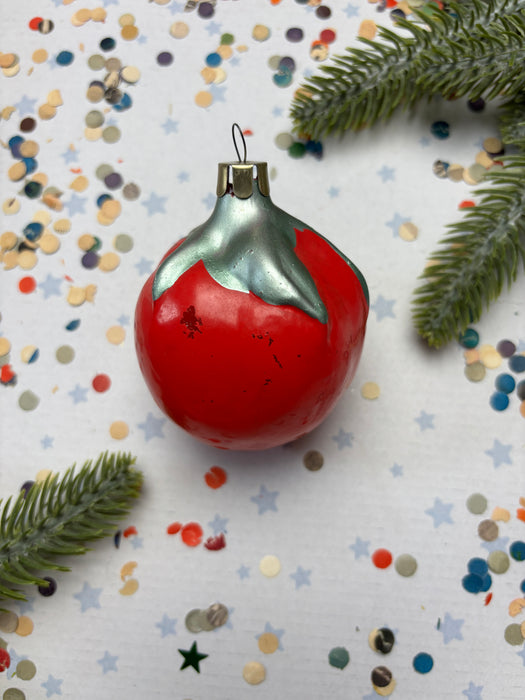 A red ornament with a silver finial is surrounded by colorful confetti and a sprig of greenery on a white background.