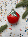 A red ornament with a silver finial is surrounded by colorful confetti and a sprig of greenery on a white background.