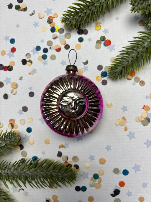 A purple glass ornament with a sun design is surrounded by confetti and pine branches on a white background.