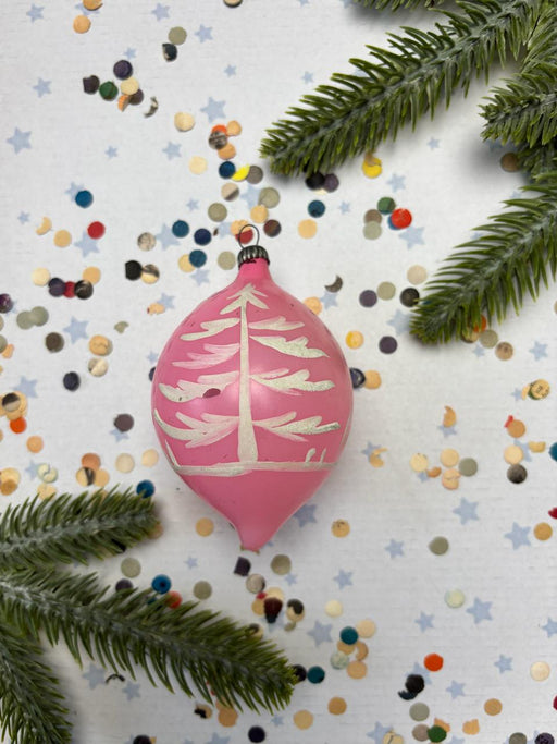 A pink Christmas ornament with a white tree design is surrounded by colorful confetti and green pine branches on a white background.