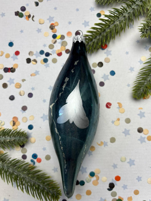 A dark blue glass ornament with a white bird design is surrounded by colorful confetti and pine branches on a white background.