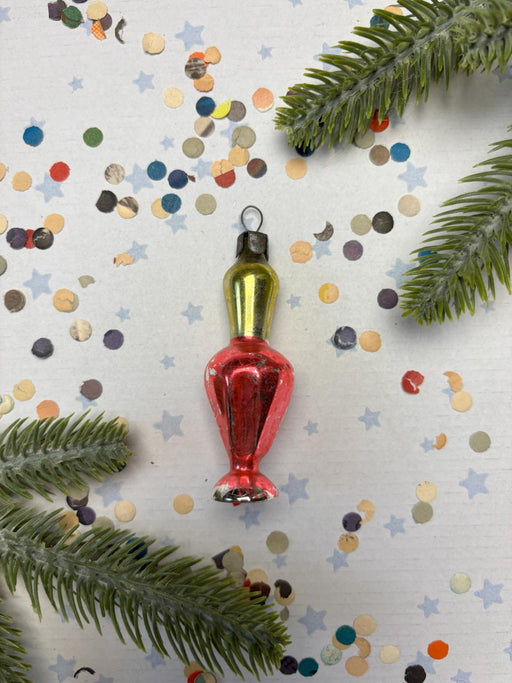 a red and yellow ornament with a green base, surrounded by confetti and pine branches.