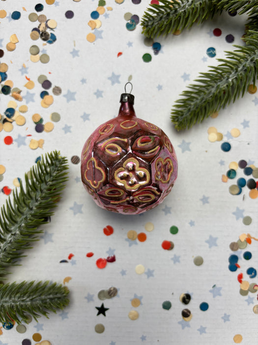 A pink ornament with gold designs sits on a white background, surrounded by scattered confetti and a sprig of greenery.